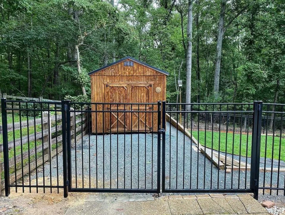 Black aluminum fence with double gate in front of a wooden storage shed on gravel.
