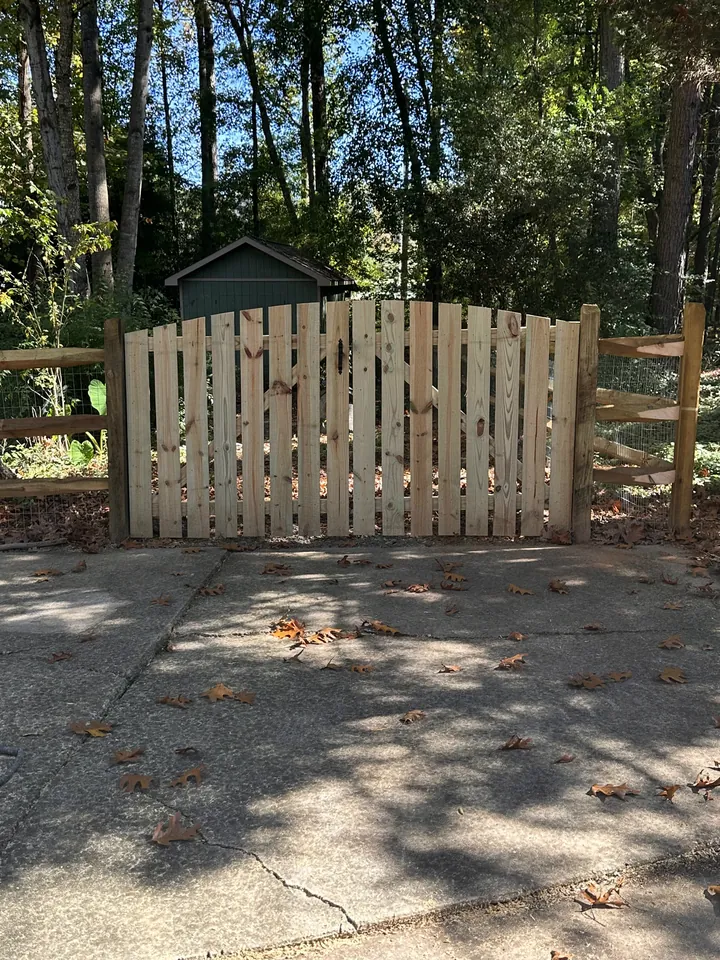 Wood picket style drive gate set in a split rail fence over a concrete driveway.